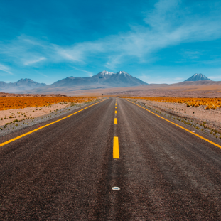 picture of a black road with yellow lines in the desert with a blue sky and mountains