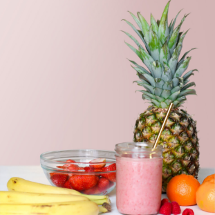  picture of a pinaple pink cup with straw, strawberries in a clear bowl, oranges, bannanas on a table with a pink background.